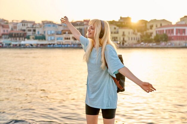 Mujer rubia feliz disfrutando en auriculares con mochila en el paseo marítimo