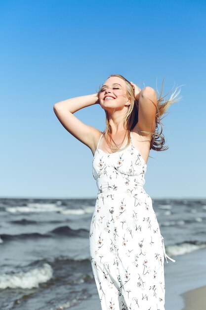 Mujer rubia feliz en la felicidad de la felicidad libre en la playa del océano de pie derecho. Retrato de una modelo femenina con vestido blanco de verano disfrutando de la naturaleza durante las vacaciones de viaje al aire libre.