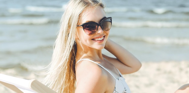 Mujer rubia feliz con gafas de sol y relajándose en una tumbona de madera en la playa del océano.
