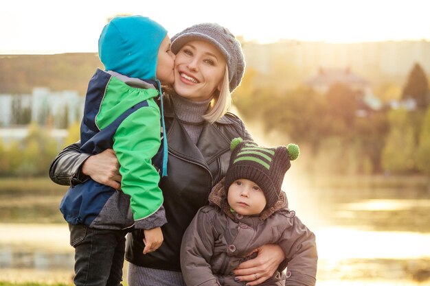 Mujer rubia feliz y hermosa con sombrero jugando a dos hijos en una chaqueta cálida sosteniéndolo en sus brazos en el parque de otoño contra el telón de fondo del lago elegante madre con un niño