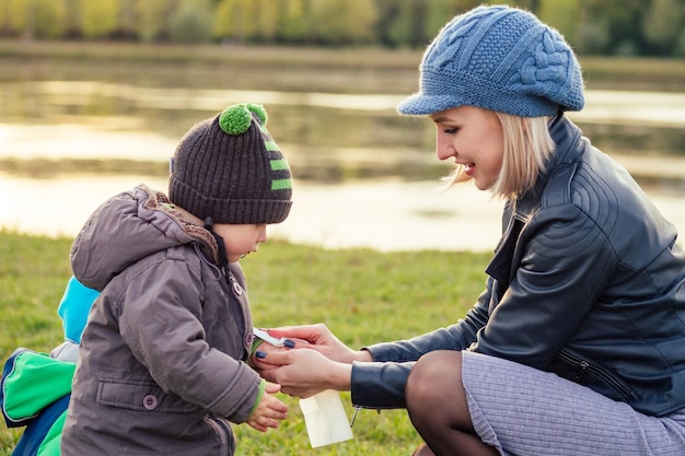 Mujer rubia feliz y hermosa con sombrero jugando a limpiarse las manos con servilletas de hijo en una chaqueta cálida en el parque de otoño con el telón de fondo del lago elegante madre con un niño