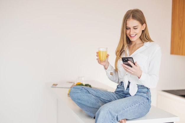 Mujer rubia feliz y sonriente sentada encima de la mesa de la cocina bebiendo jugo de naranja y hablando por teléfono