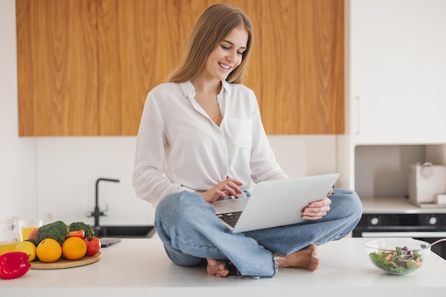 Mujer rubia feliz y sonriente sentada encima de la mesa en la cocina y buscando recetas en Internet en la computadora portátil