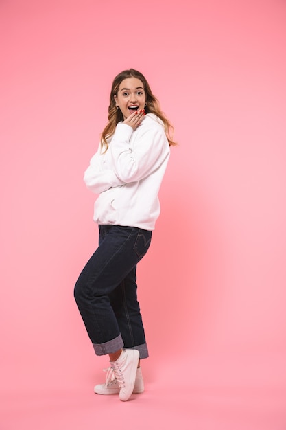 Mujer rubia feliz sorprendida de longitud completa vestida con ropa casual se regocija y mirando al frente sobre la pared rosa