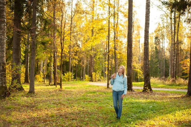 Foto una mujer rubia feliz tranquila camina en el bosque de otoño con un vaso de café en sus manos. paseo de otoño. un estilo de vida saludable.