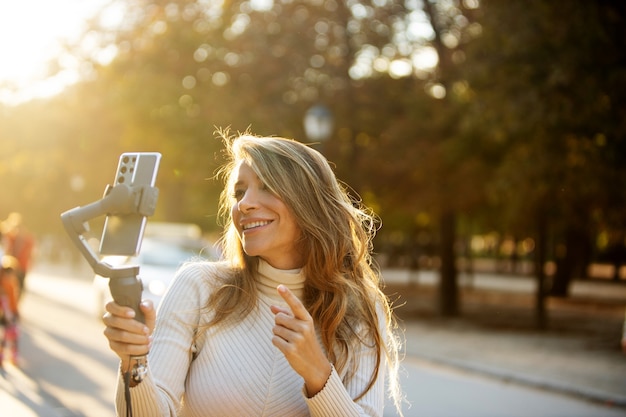 Mujer rubia filmando a sí misma al aire libre con un teléfono celular y estabilizador en el sol vespertino