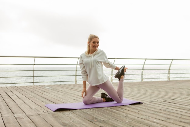 Una mujer rubia en forma practicando estiramientos de piernas antes de hacer ejercicio al aire libre. Niña vestida con capucha blanca entrena en la playa mientras se sienta en la alfombra. Estilo de vida saludable, deportes y fitness.