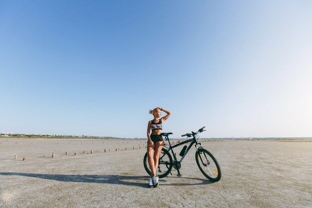 La mujer rubia fuerte con un traje multicolor se encuentra cerca de una bicicleta en una zona desértica y mira al sol. Concepto de fitness.