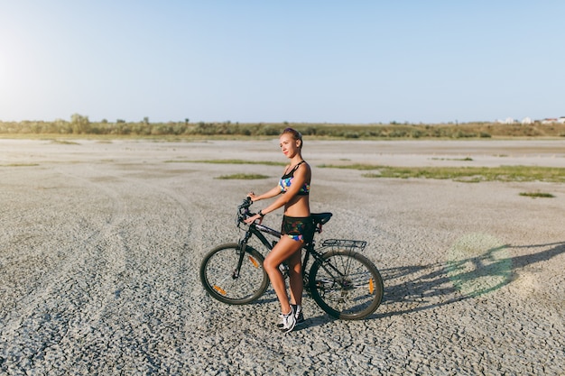 La mujer rubia fuerte con un traje multicolor se encuentra cerca de una bicicleta en una zona desértica y mira al sol. Concepto de fitness.