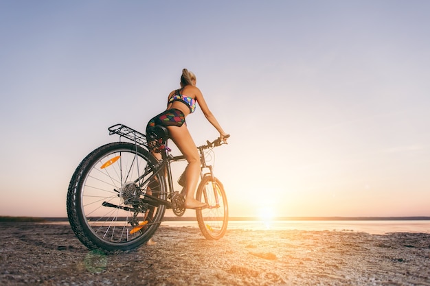 La mujer rubia fuerte con un traje multicolor se sienta en una bicicleta en una zona desértica cerca del agua y mira al sol. Concepto de fitness. Vista trasera