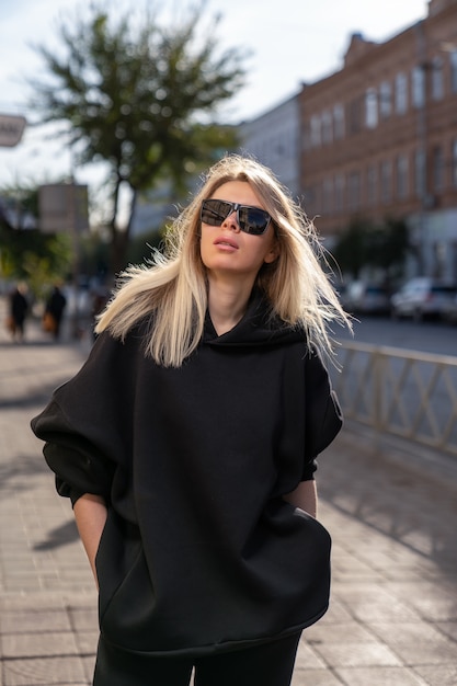 Mujer rubia con gafas de sol con una sudadera con capucha negra camina por la ciudad