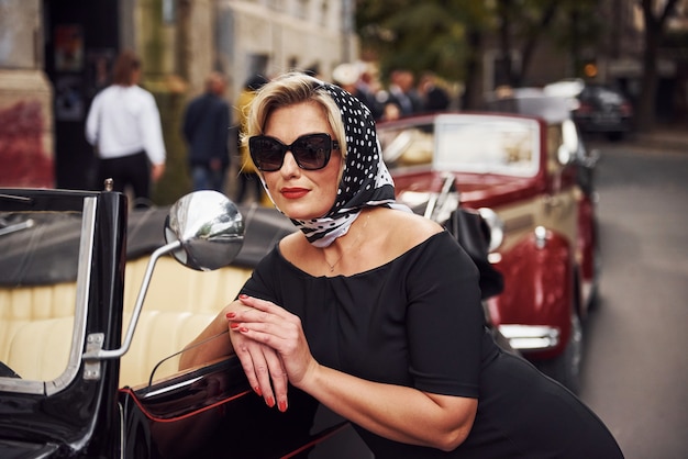 Mujer rubia con gafas de sol y vestido negro apoyado en el viejo coche clásico de época.