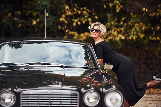 Mujer rubia con gafas de sol y vestido negro cerca del viejo coche clásico vintage.