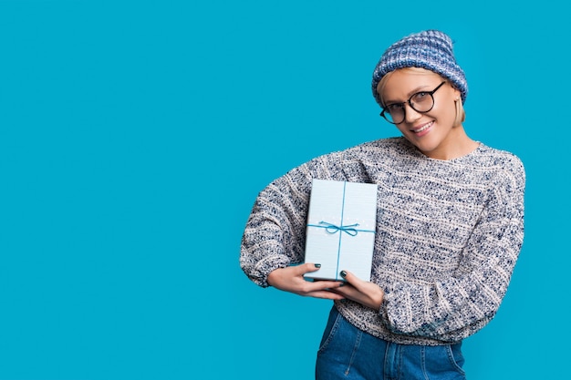 Mujer rubia con gafas y sombrero sostiene un regalo y sonríe a la cámara mientras promociona algo en una pared azul del estudio