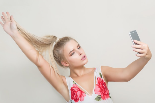 Mujer rubia glamour tirando de su cabello y haciendo selfie en teléfono móvil contra un fondo gris