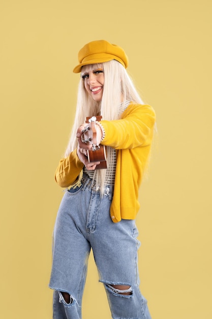 Mujer rubia con gorra y chaqueta amarilla sonriendo y tocando el ukelele con fondo amarillo