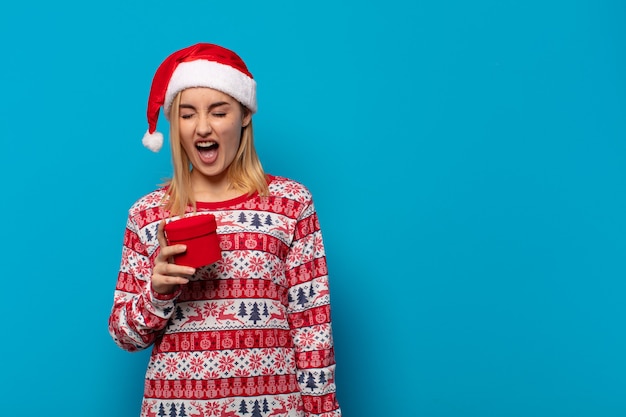 Mujer rubia con gorro de Papá Noel gritando agresivamente, luciendo muy enojado, frustrado, indignado o molesto, gritando no