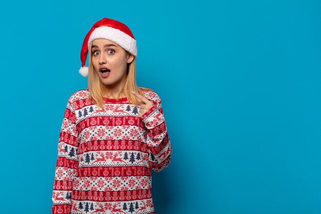 Mujer rubia con gorro de Papá Noel mirando consternado y sorprendido con la boca abierta, apuntando a sí mismo