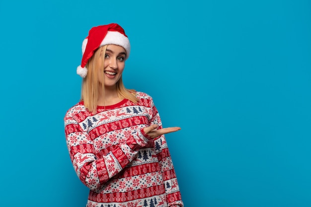 Mujer rubia con gorro de Papá Noel sonriendo alegremente, sintiéndose feliz y mostrando un concepto en el espacio de la copia con la palma de la mano