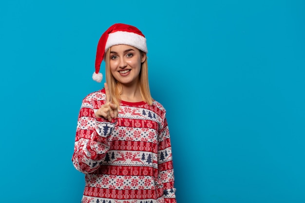 Mujer rubia con gorro de Papá Noel sonriendo y mirando amistosamente, mostrando el número uno o el primero con la mano hacia adelante, contando hacia atrás