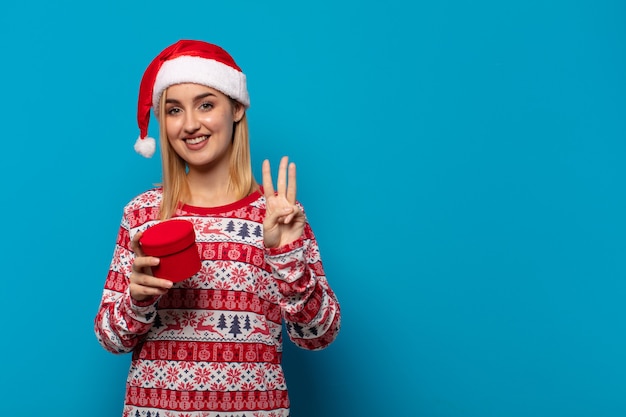 Mujer rubia con gorro de Papá Noel sonriendo y mirando amistosamente, mostrando el número tres o tercero con la mano hacia adelante, contando hacia atrás