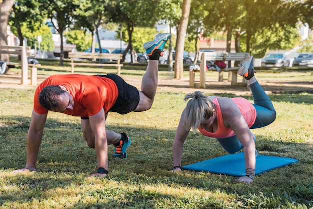 Mujer rubia haciendo ejercicios en una colchoneta en un parque con un entrenador personal a su lado