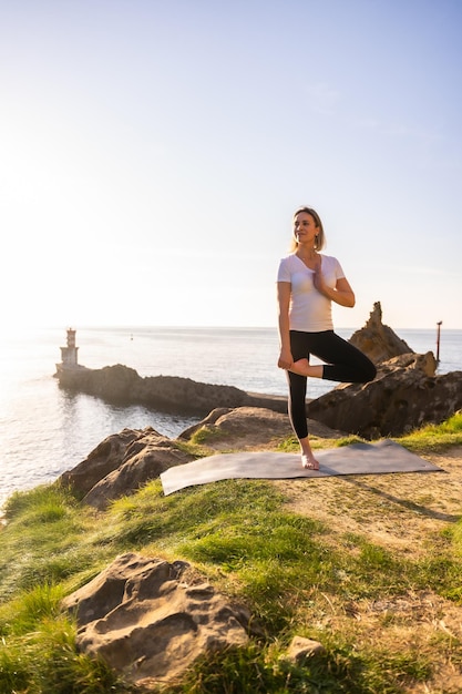 Una mujer rubia haciendo ejercicios de yoga en la naturaleza junto al mar estirándose junto a un faro vida sana y naturista pilates al aire libre