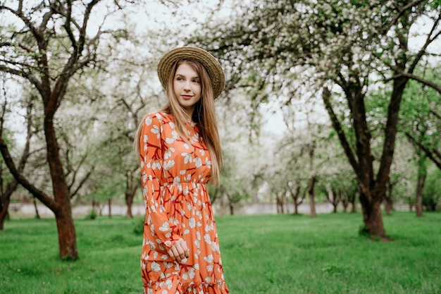 Mujer rubia hermosa joven en jardín floreciente. Árboles de primavera en flor. Vestido naranja y sombrero de paja.