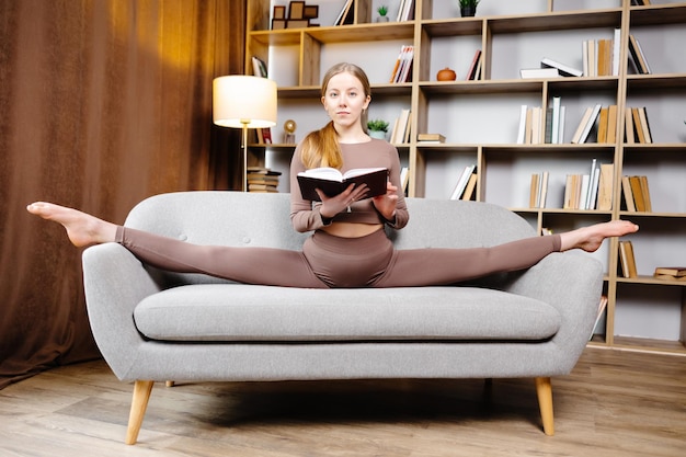 Mujer rubia hermosa joven en polainas marrones y top sentado en un hilo en casa en el sofá La niña está leyendo un libro sentado en el sofá en una posición inusual