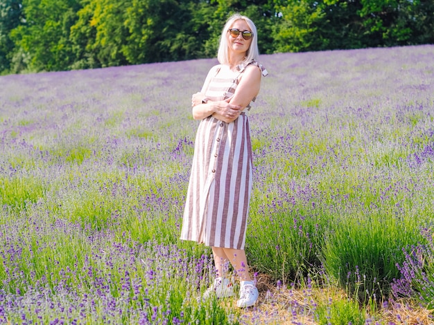 La mujer rubia hermosa en vestido se sienta entre los campos de la lavanda en Provence. Campos de lavanda violeta que florece en la luz del sol de verano. Mar de Lila Flores paisaje. Ramo de flores perfumadas de la Provenza francesa
