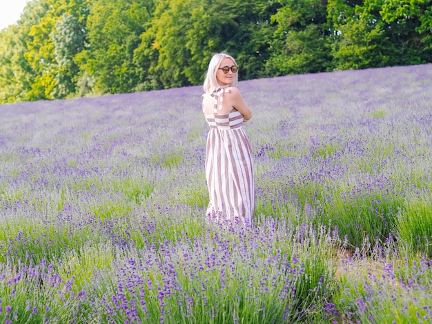 La mujer rubia hermosa en vestido se sienta entre los campos de la lavanda en Provence. Campos de lavanda violeta que florece en la luz del sol de verano. Mar de Lila Flores paisaje. Ramo de flores perfumadas de la Provenza francesa