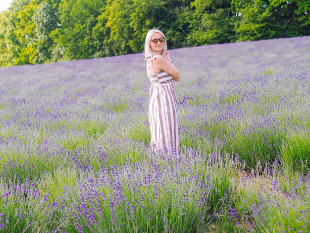 La mujer rubia hermosa en vestido se sienta entre los campos de la lavanda en Provence. Campos de lavanda violeta que florece en la luz del sol de verano. Mar de Lila Flores paisaje. Ramo de flores perfumadas de la Provenza francesa