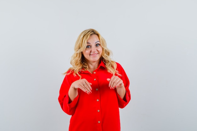 Mujer rubia invitando a venir en blusa roja y mirando feliz, vista frontal.