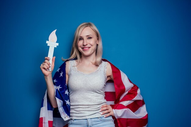 Mujer rubia en jeans sosteniendo la bandera estadounidense con corona de papel y antorcha Estatua de la libertad sobre un fondo azul en el estudio Concepto de celebración del día de la independencia del 4 de julio