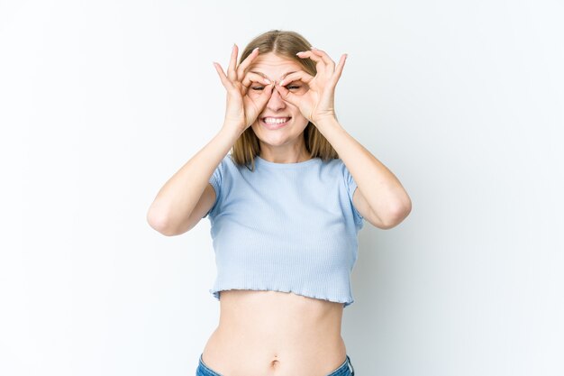 Mujer rubia joven aislada en la pared blanca emocionada manteniendo el gesto ok en el ojo.