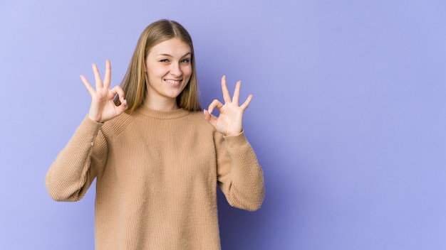 Mujer rubia joven aislada en la pared púrpura alegre y confiada que muestra el gesto aceptable.