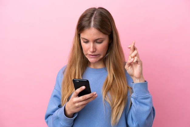 Mujer rubia joven aislada sobre fondo rosa a través de teléfono móvil con los dedos cruzados