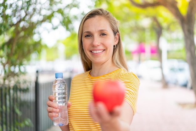 Mujer rubia joven al aire libre con una manzana y una botella de agua