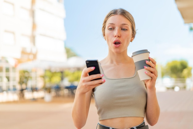 Mujer rubia joven al aire libre usando teléfono móvil y sosteniendo un café con expresión sorprendida