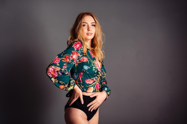 Mujer rubia joven apoyada en una cadera en una blusa con un estampado floral sobre un fondo gris