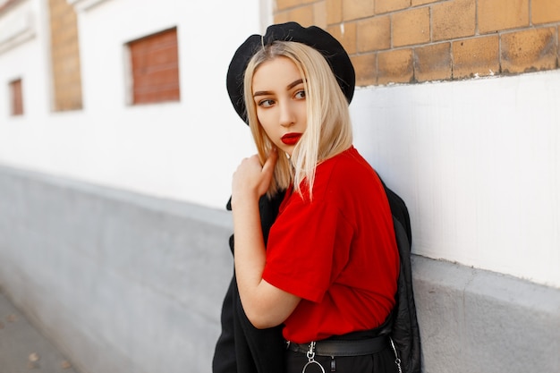 La mujer rubia joven bastante moderna de moda con los labios atractivos en un abrigo negro de moda con una camiseta roja con estilo en una boina elegante está de pie en la ciudad cerca de un edificio de época