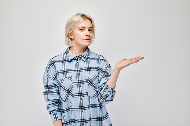 Mujer rubia joven en casual sosteniendo algo en la mano demostrando espacio vacío para producto o texto aislado en fondo de estudio blanco