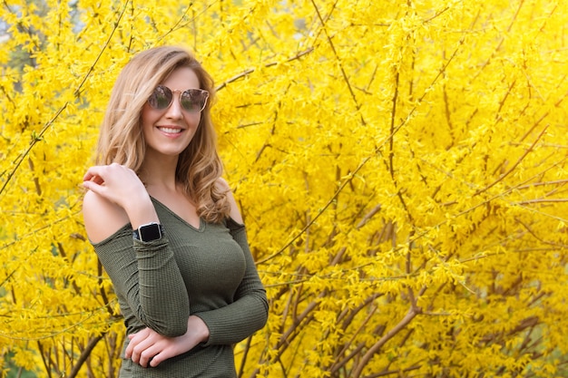 La mujer rubia joven cerca de árboles amarillos florecientes en primavera estaciona el día soleado.