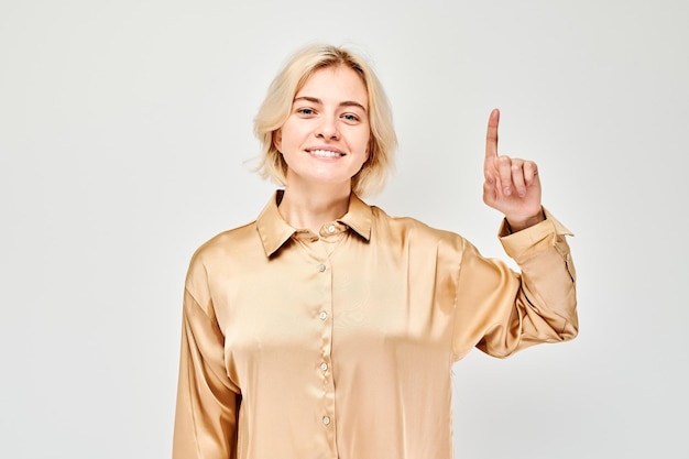 Mujer rubia joven con el dedo apuntando hacia arriba demostrando el espacio vacío para el producto o el texto aislado en el fondo blanco del estudio