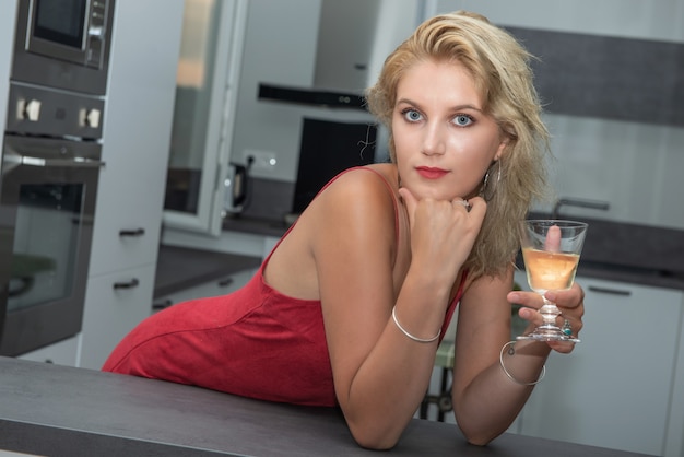 Mujer rubia joven hermosa con el vestido blanco que bebe el vino blanco en la cocina