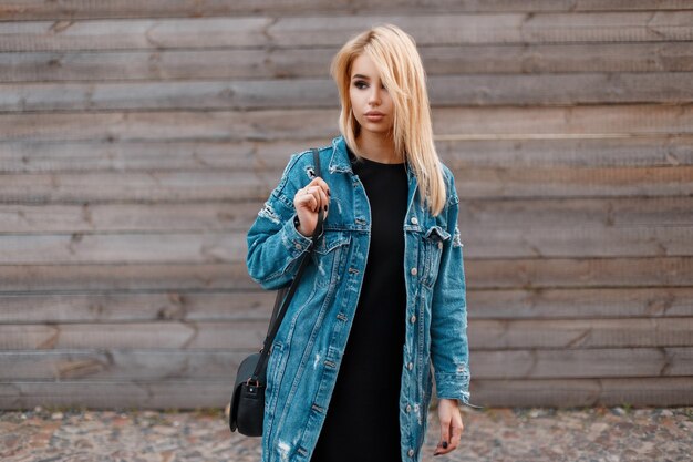 Mujer rubia joven de moda moderna con bolso negro de moda en una chaqueta de mezclilla con estilo largo con un vestido negro está de pie cerca de la pared de madera vintage al aire libre