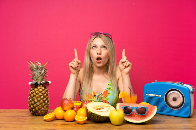 Foto mujer rubia joven con muchas frutas que señala con el dedo índice una gran idea