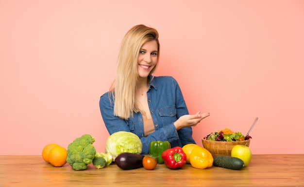 Mujer rubia joven con muchas verduras que presenta una idea mientras que mira sonriente hacia