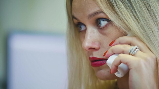 Mujer rubia joven en la oficina hablando por teléfono frente a la computadora muy de cerca