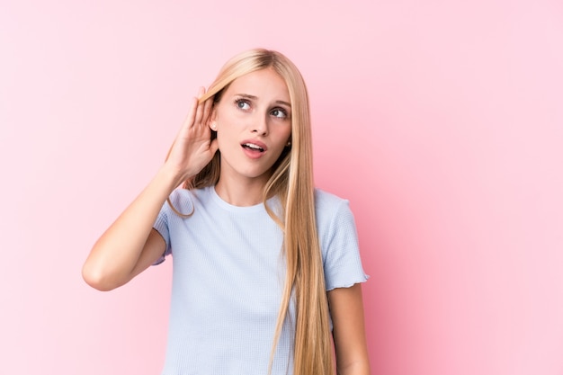 Mujer rubia joven en la pared rosada que intenta escuchar un chisme.